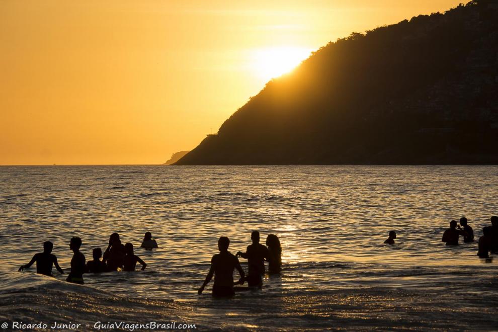 Imagem de pessoas no mar e ao fundo lindo por do sol na Praia de Ipanema.